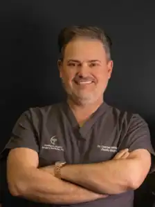 Smiling man in gray scrubs with arms crossed, wearing a gold watch, against a dark background.